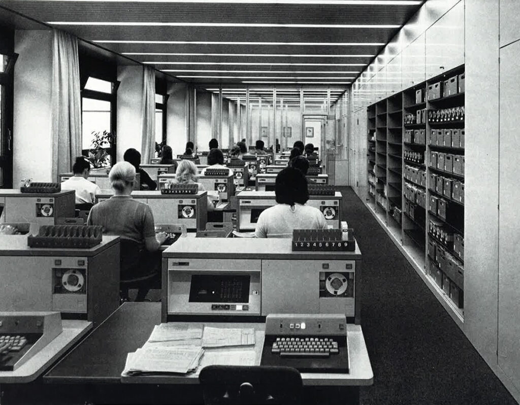 Großraumbüro Anfang der 1970er Jahre mit mehreren Personen an frühen Datenverarbeitungsgeräten, Reihen von Arbeitsplätzen mit Monitoren und Tastaturen sowie Aktenregalen im Hintergrund.