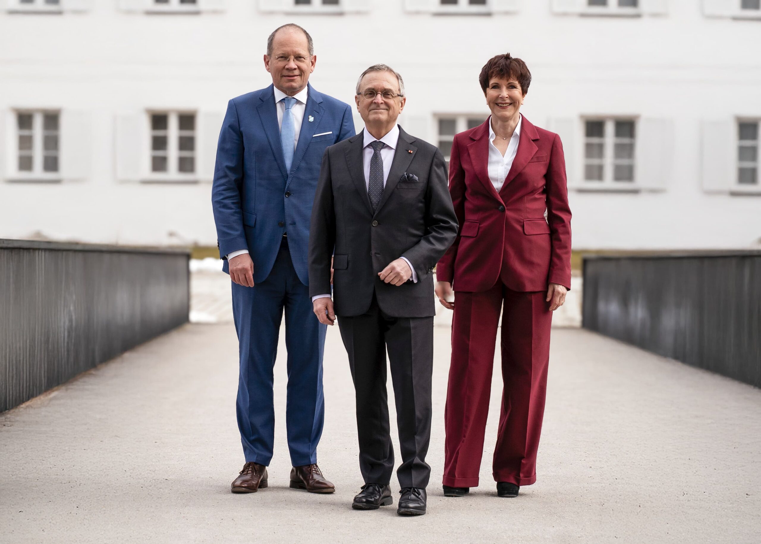Drei Personen in formeller Kleidung stehen vor einem Gebäude, Gruppenporträt des Präsidiums vor neutralem Hintergrund.